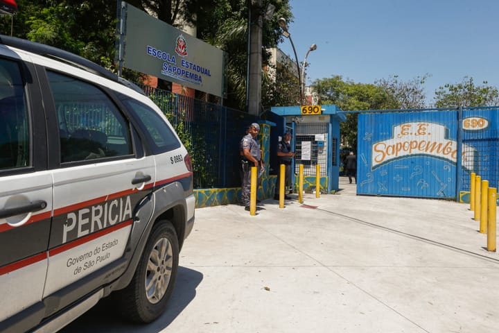 Dois policiais estão perto do portão, que é azul e etiquetado com o nome da escola em uma área ensolarada com árvores.