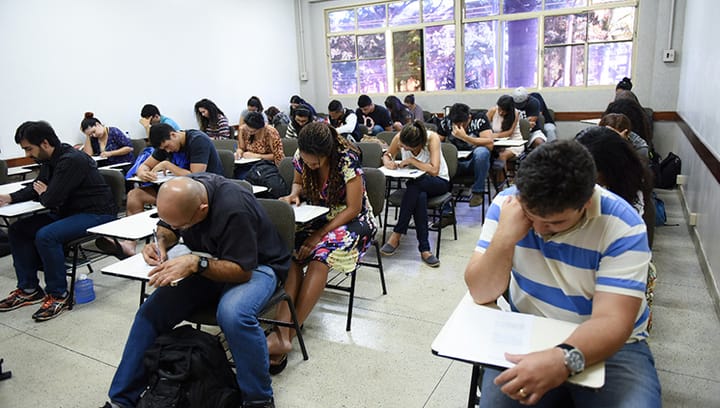 Alunos sentados em fileiras em uma sala de aula se concentram na escrita durante uma prova.