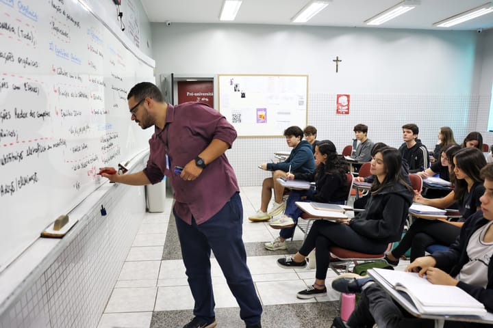 Um professor escreve em um quadro branco em uma sala de aula enquanto os alunos sentam em fileiras, ouvindo e tomando notas.