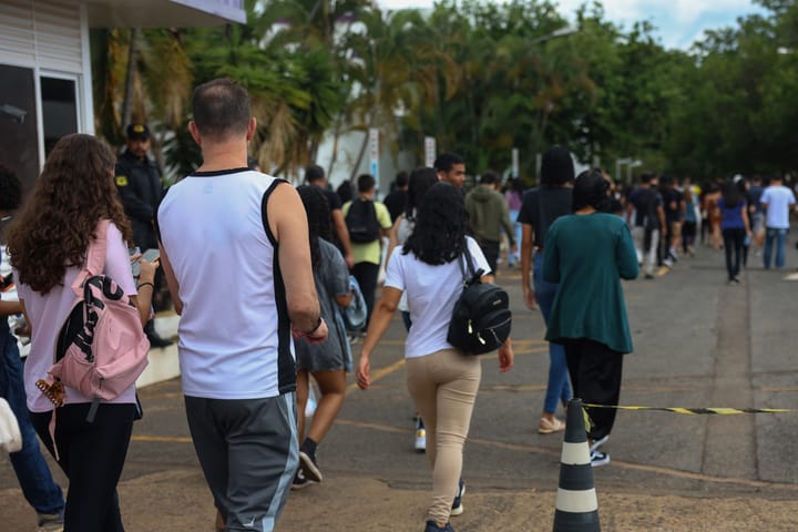 Uma multidão de pessoas anda em fila do lado de fora em um caminho pavimentado, com cones de trânsito marcando a rota.